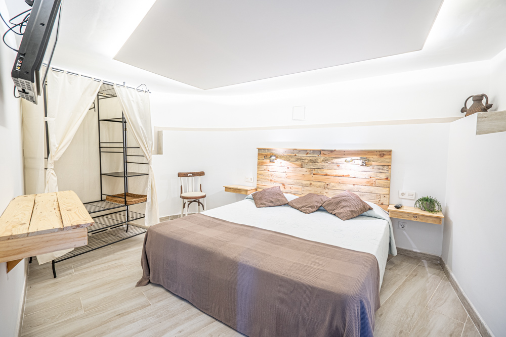 Chambre avec tête de lit en bois rustique, tables de chevet flottantes, éclairage indirect au plafond et un dressing ouvert avec rideaux blancs.