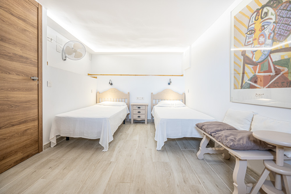 Chambre lumineuse avec deux lits simples, sol en grès cérame imitation bois, éclairage LED indirect au plafond et ventilateur mural.