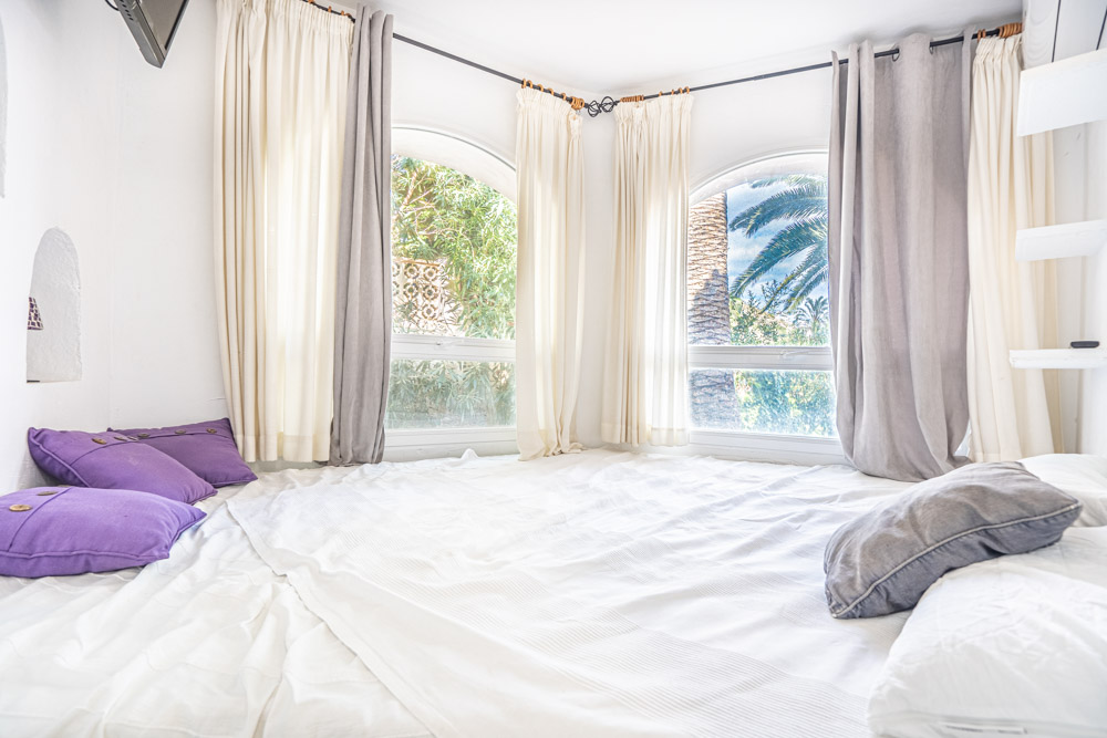 Chambre lumineuse avec fenêtres en arc offrant une vue sur le jardin méditerranéen. Décoration épurée en blanc avec des touches de violet et climatisation.
