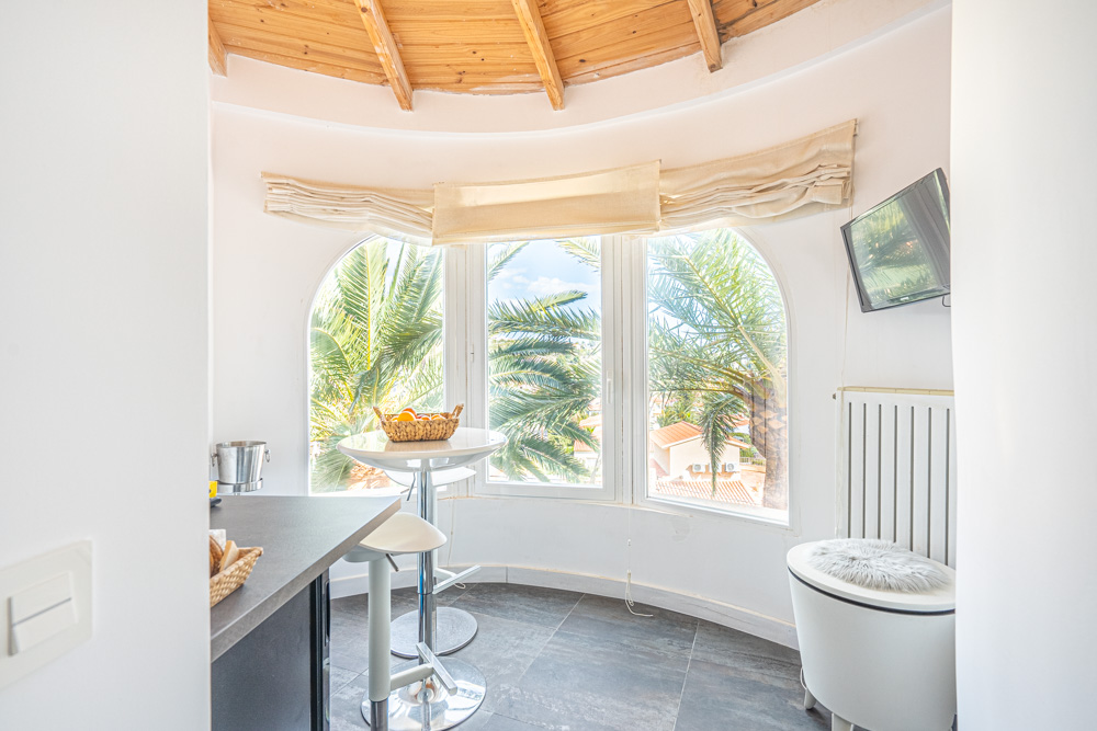 Coin petit-déjeuner avec fenêtres en plein cintre offrant une vue sur les palmiers, plafond en poutres de bois et table haute avec tabourets.