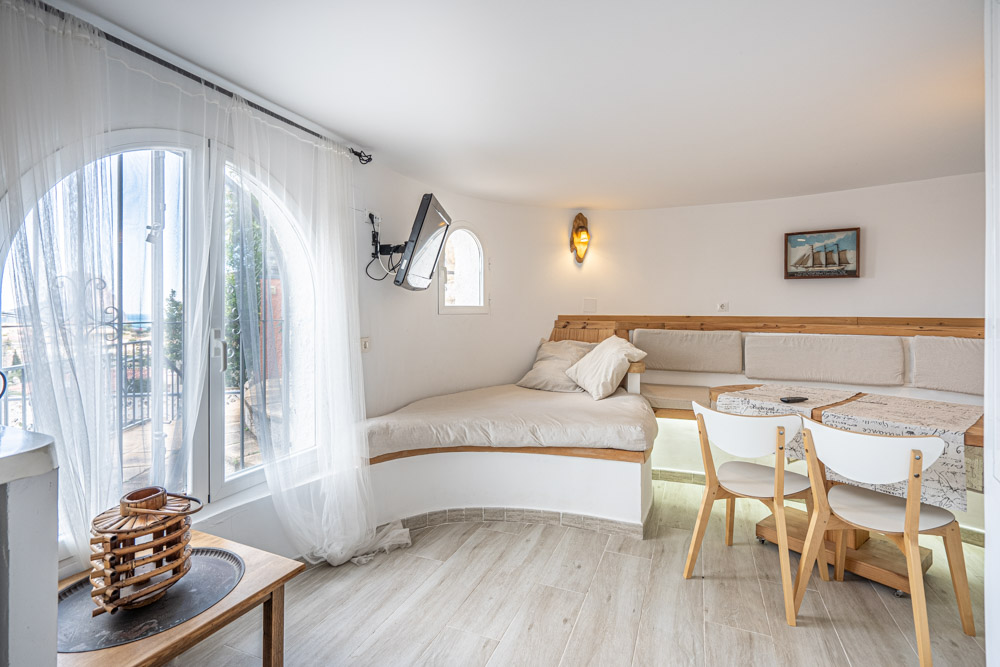 Coin salon avec banquette en bois intégrée, table à manger et chaises blanches. La pièce dispose d'une fenêtre en arc et d'un sol en grès cérame imitation bois.