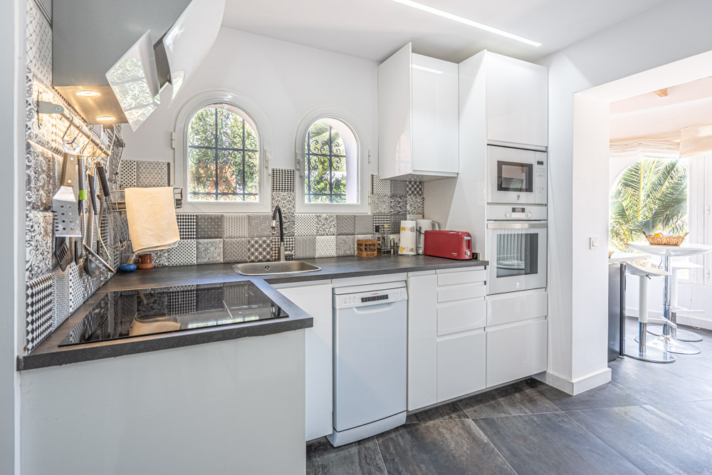 Cuisine moderne avec des meubles blancs brillants, un plan de travail sombre et une crédence en carreaux de ciment. Fenêtres cintrées et électroménagers encastrés inclus.