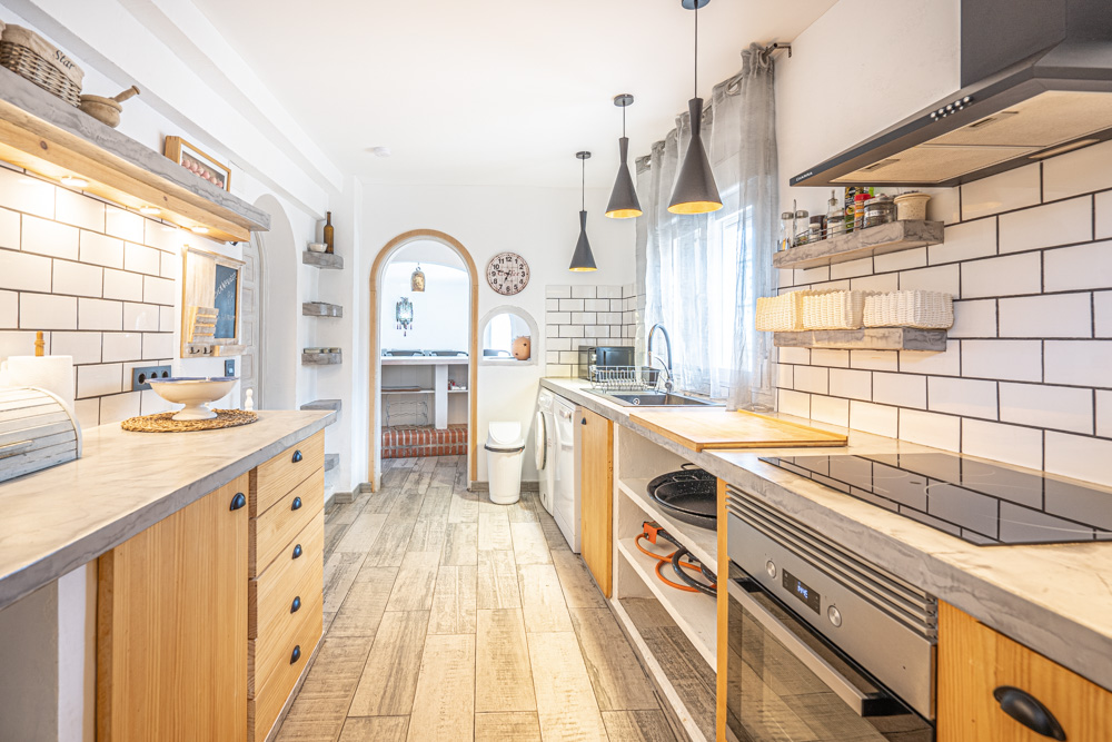 Cuisine rustique avec plans de travail gris, meubles en bois et carrelage métro blanc. Elle comprend des appareils intégrés, des suspensions noires et une arche vers la salle à manger.