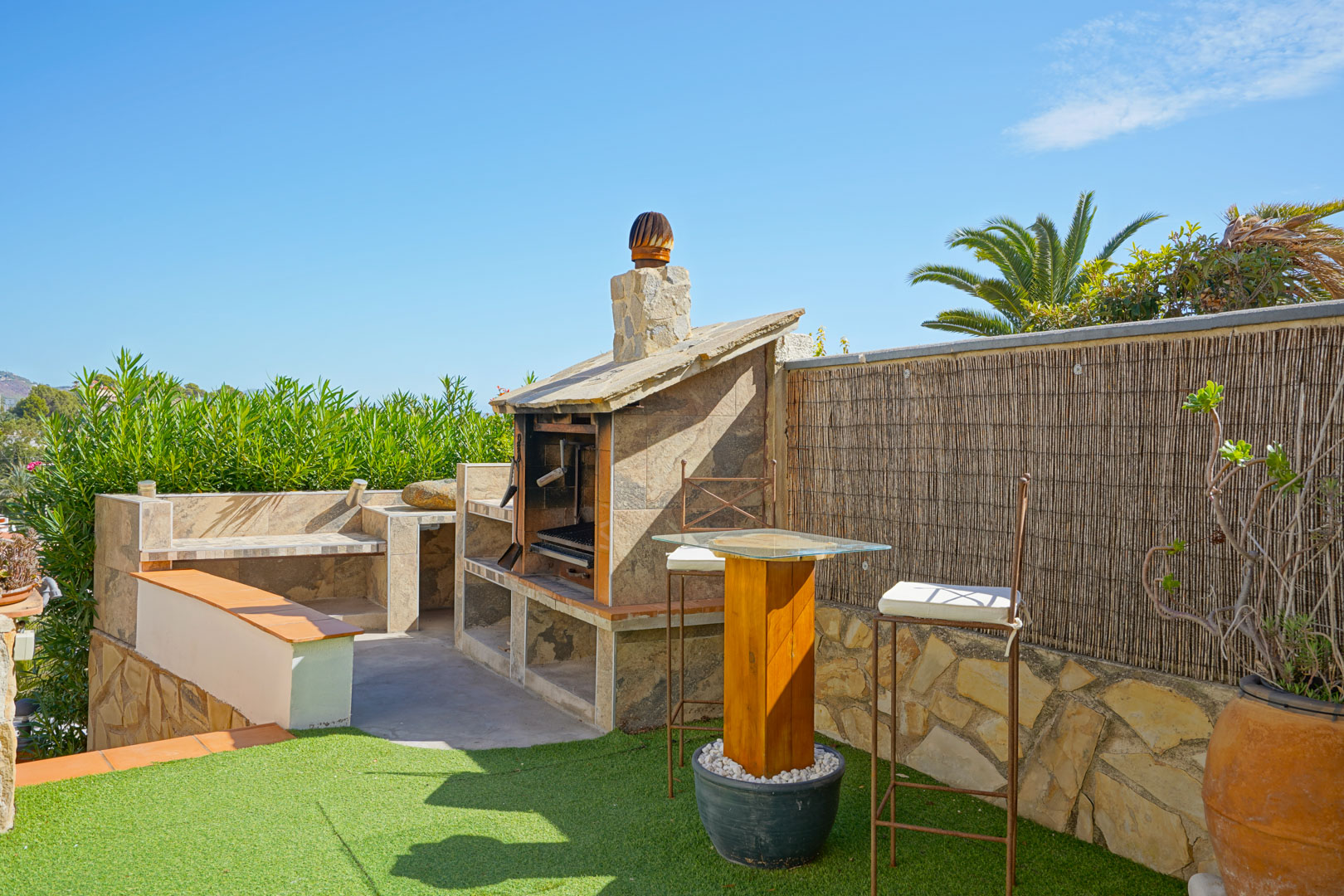 Espace barbecue extérieur en pierre avec plans de travail carrelés, mange-debout en bois et tabourets sur gazon synthétique.
