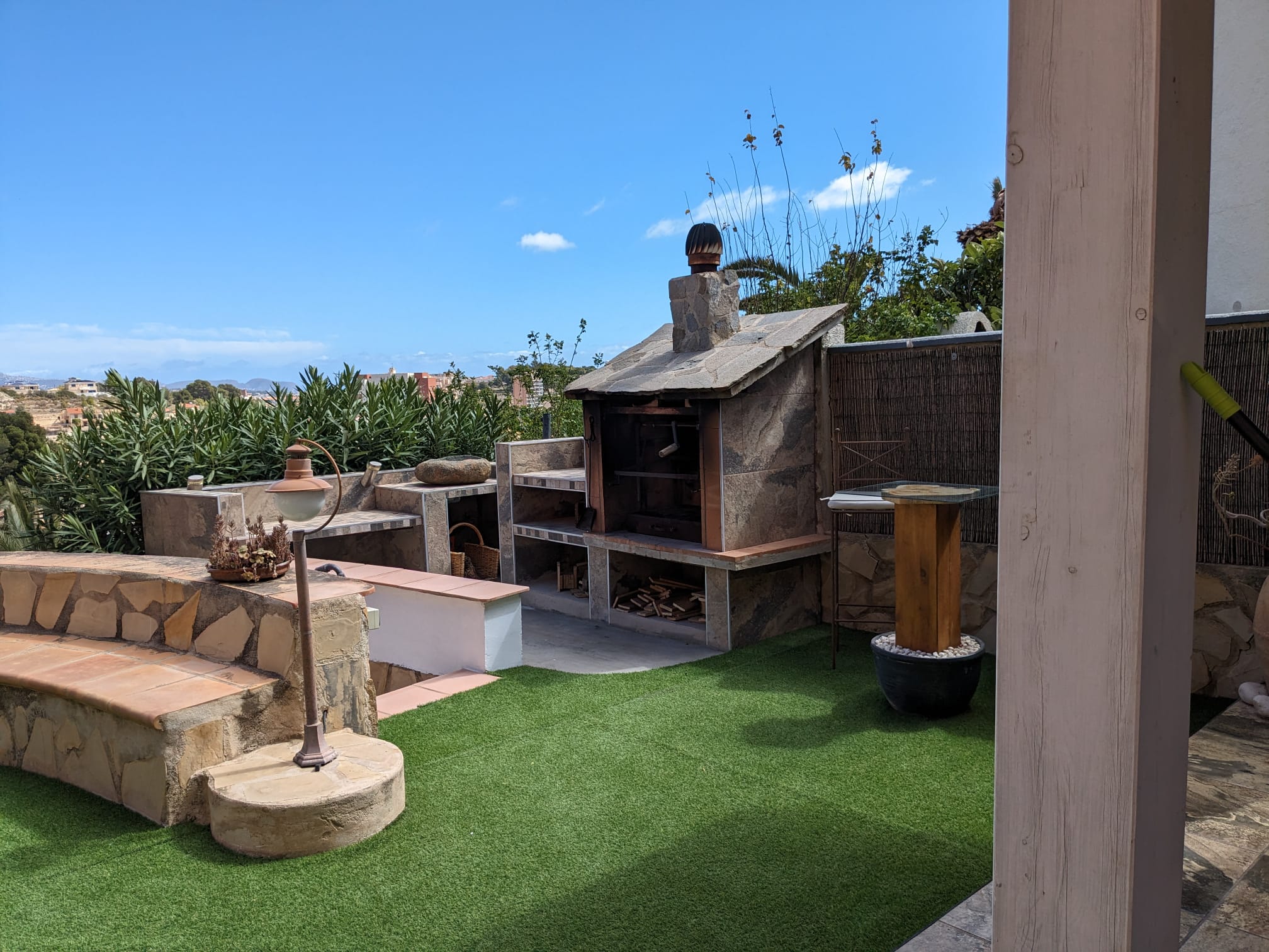 Espace barbecue extérieur en pierre naturelle avec plans de travail intégrés, banc maçonné et pelouse artificielle sous un ciel azur.