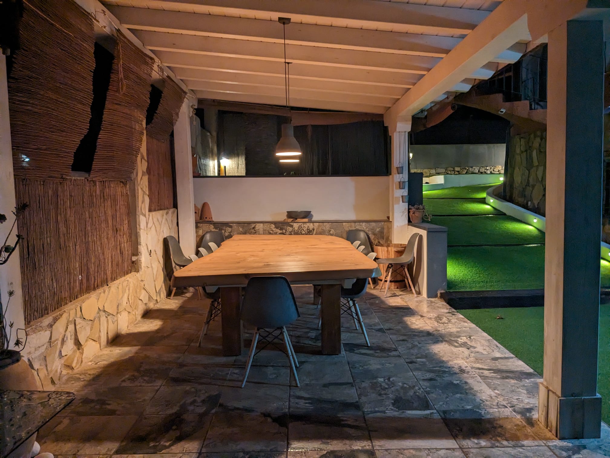 Espace dînatoire extérieur couvert avec table en bois massif, murs en pierre et éclairage d'ambiance nocturne donnant sur l'allée du jardin.