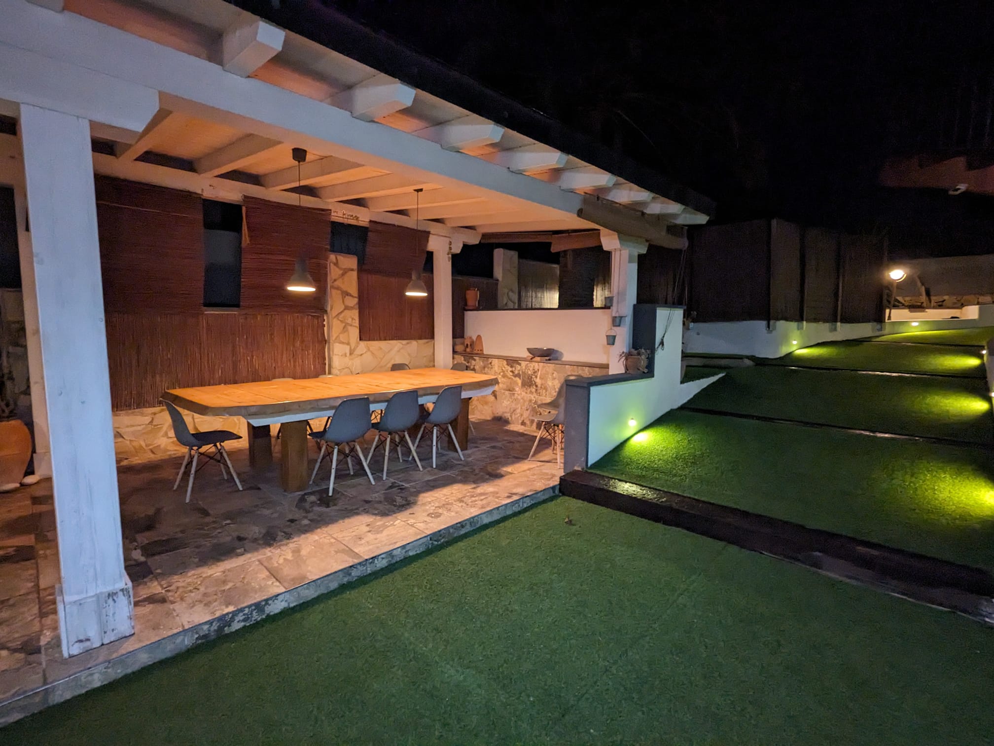 Espace dînatoire extérieur sous une pergola en bois blanc avec table massive, chaises modernes et sol en pierre, bordé d'un jardin éclairé par des LED.