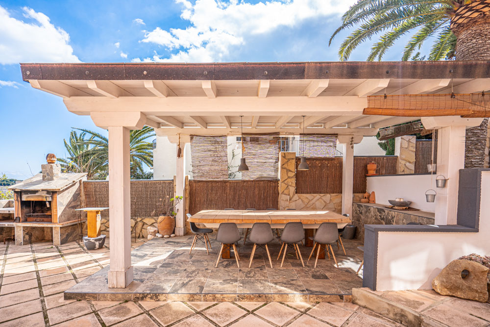 Espace dînatoire extérieur sous une pergola en bois blanc, comprenant une grande table en bois massif, des chaises modernes et un barbecue en pierre naturelle.