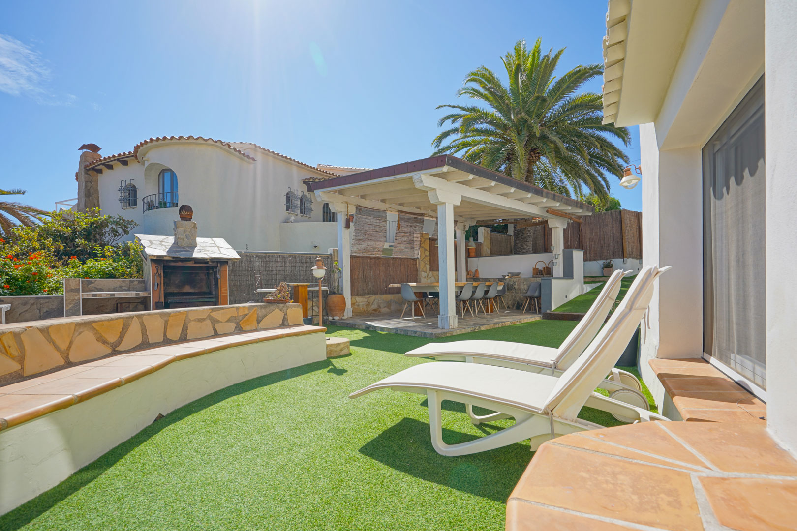 Espace extérieur avec gazon artificiel, chaises longues blanches et une pergola en bois abritant un coin repas à côté du barbecue en pierre.