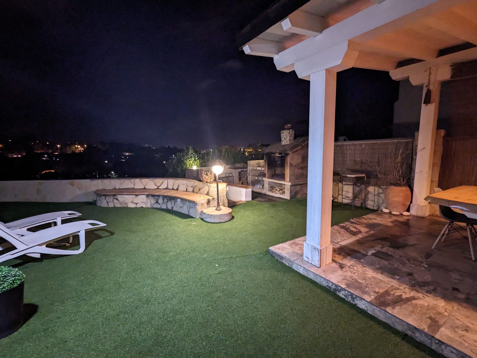 Espace extérieur nocturne comprenant un barbecue en pierre, un banc incurvé en maçonnerie et une pergola en bois éclairée à côté d'une zone en gazon synthétique.