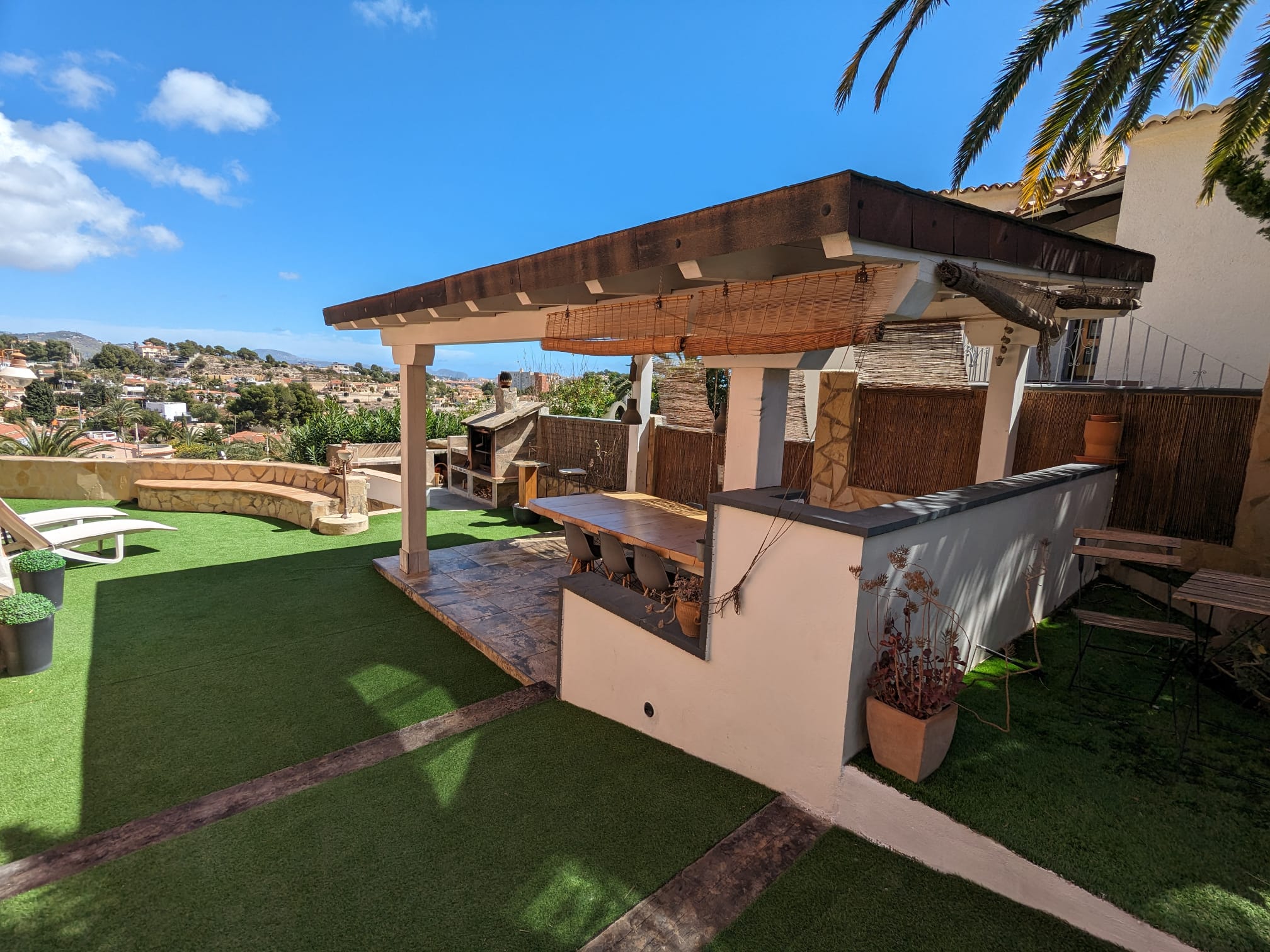 Espace repas extérieur sous une pergola en bois avec barbecue en pierre, sol en carrelage et pelouse synthétique offrant une vue sur les environs de Calpe.