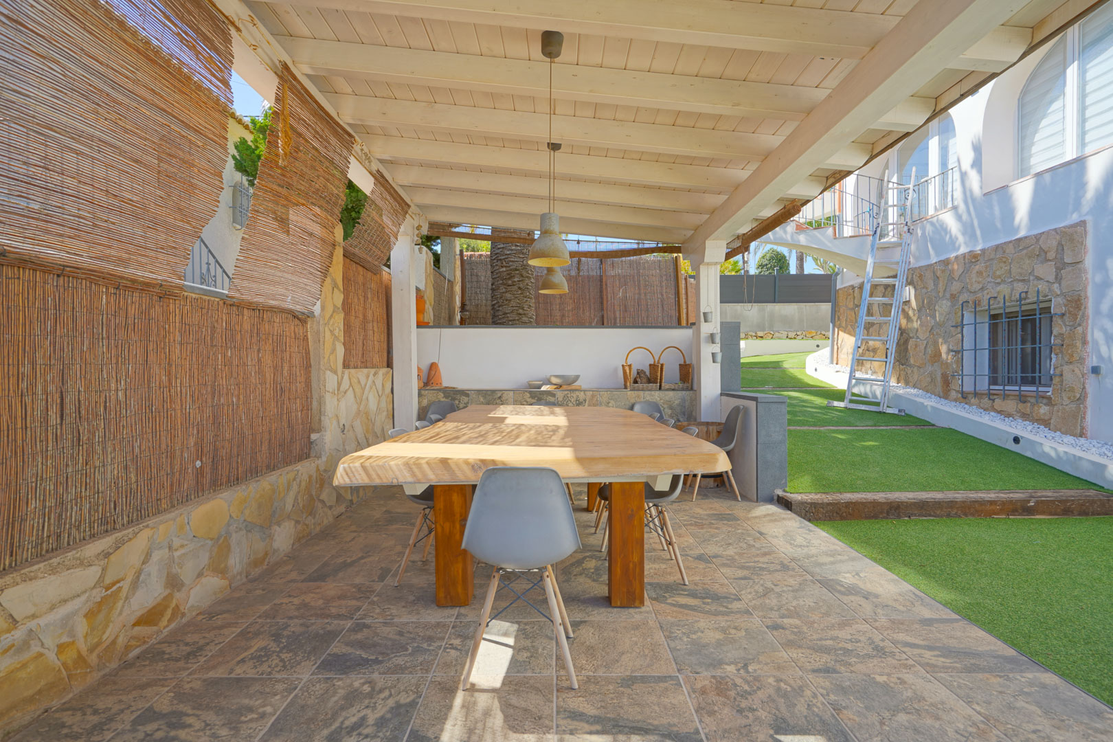 Espace repas extérieur sous une pergola en bois blanc, avec une table en bois massif, un sol en grès cérame et des murs en pierre naturelle.