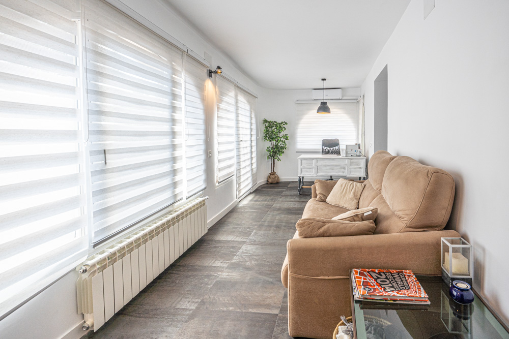 Naya lumineuse avec sol en grès cérame foncé, canapé beige et coin bureau. Équipée d'un radiateur, de la climatisation et de grandes fenêtres avec stores zébrés blancs.