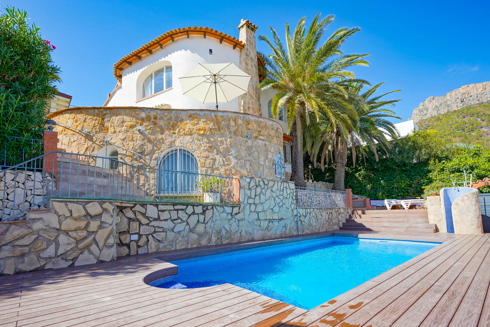 Piscine privée avec terrasse en bois et murs en pierre naturelle, entourée de palmiers et offrant une vue sur les montagnes environnantes.