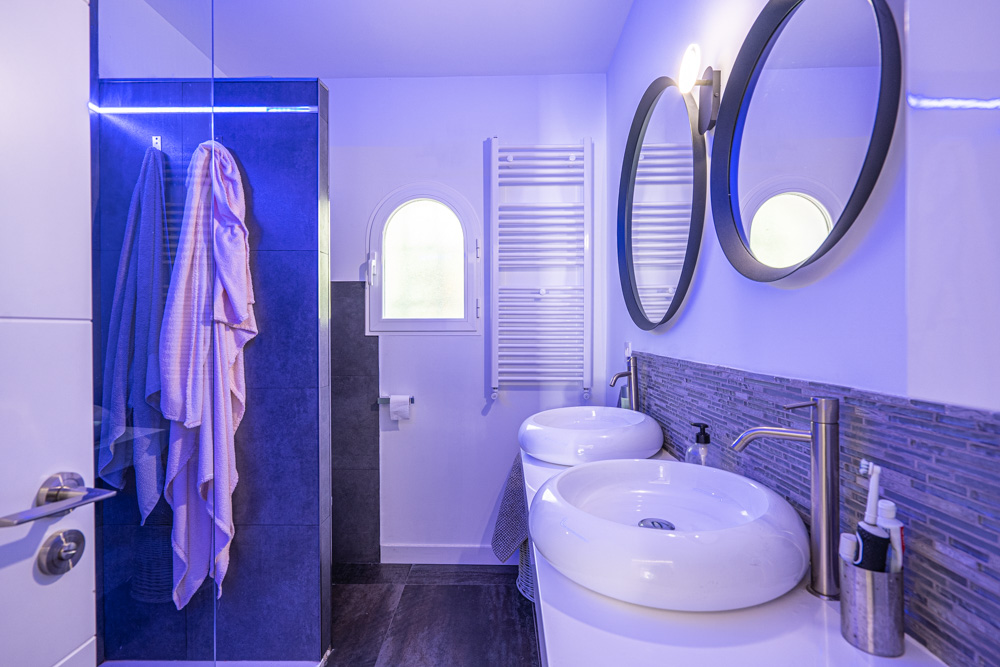 Salle de bain moderne avec double vasque circulaire, miroirs ronds et douche à l'italienne dotée d'un éclairage LED bleu intégré.