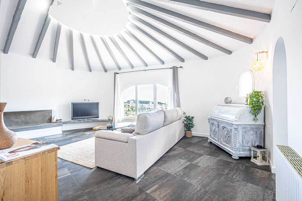 Salon circulaire avec plafond à poutres apparentes rayonnantes, grande fenêtre cintrée offrant une vue dégagée et sol en grès cérame foncé.