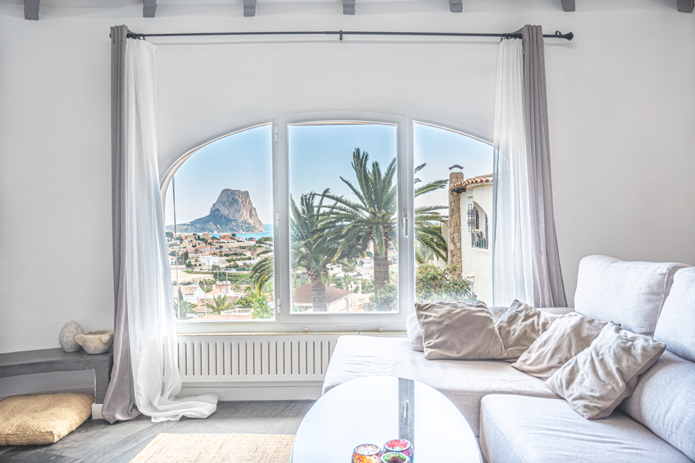 Salon lumineux doté d'une grande fenêtre en arche offrant une vue imprenable sur le Peñón de Ifach et la mer, avec des poutres apparentes au plafond.