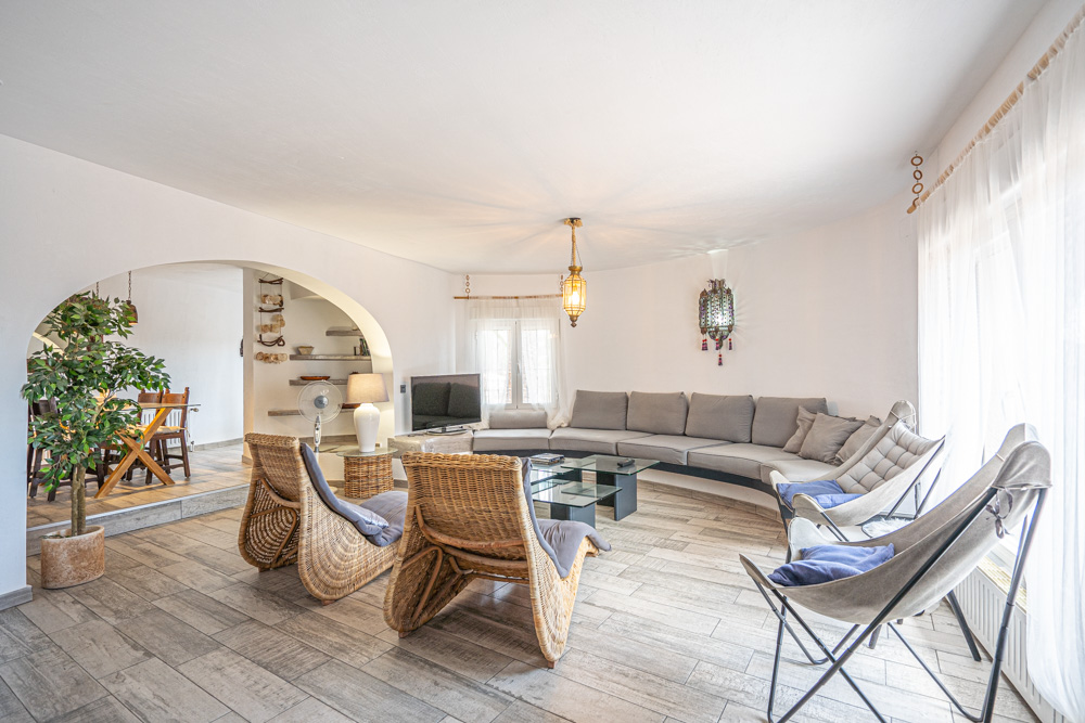 Salon spacieux avec canapé incurvé intégré, fauteuils en osier et sol en grès cérame imitation bois. Une arche élégante sépare la zone de détente de la salle à manger.
