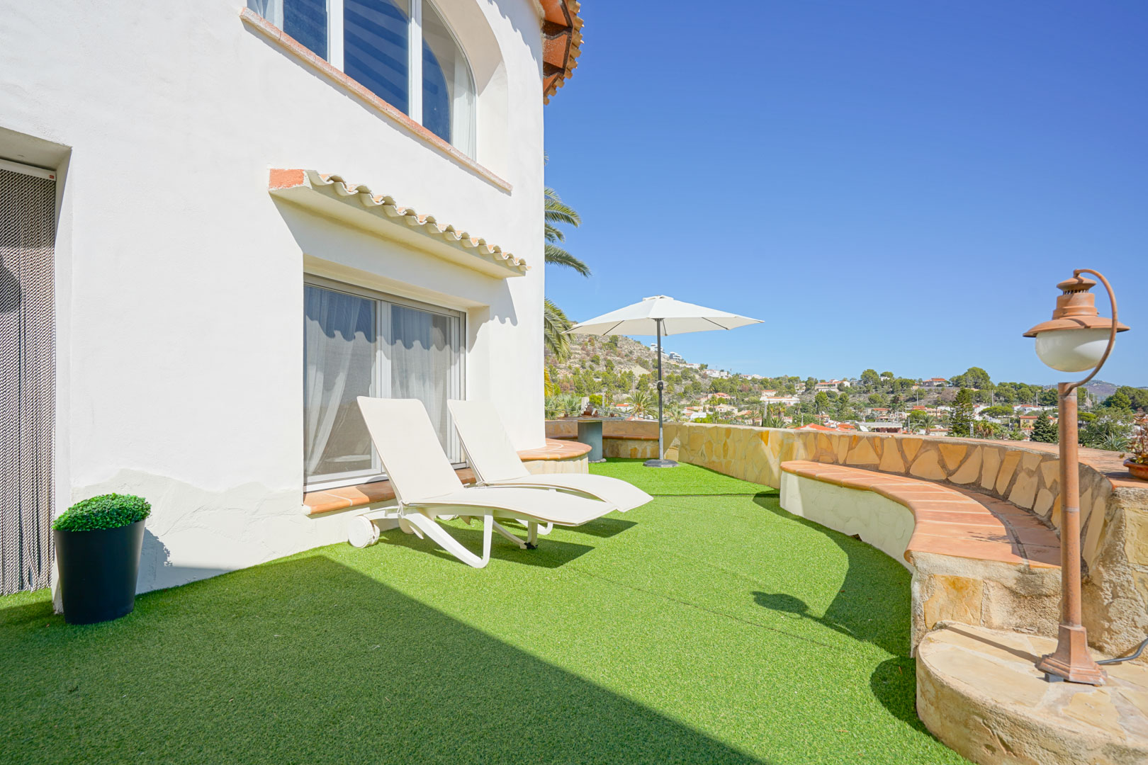 Terrasse avec gazon artificiel, chaises longues blanches et muret en pierre avec banc intégré offrant une vue panoramique sur les environs de Calpe.