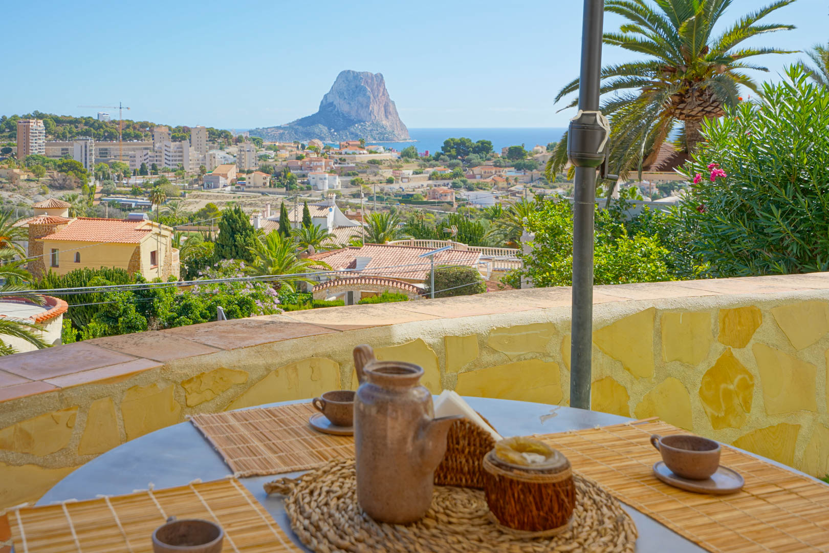 Terrasse extérieure avec coin repas offrant une vue panoramique sur le Peñón de Ifach et la mer, bordée de murs en pierre naturelle et de végétation.