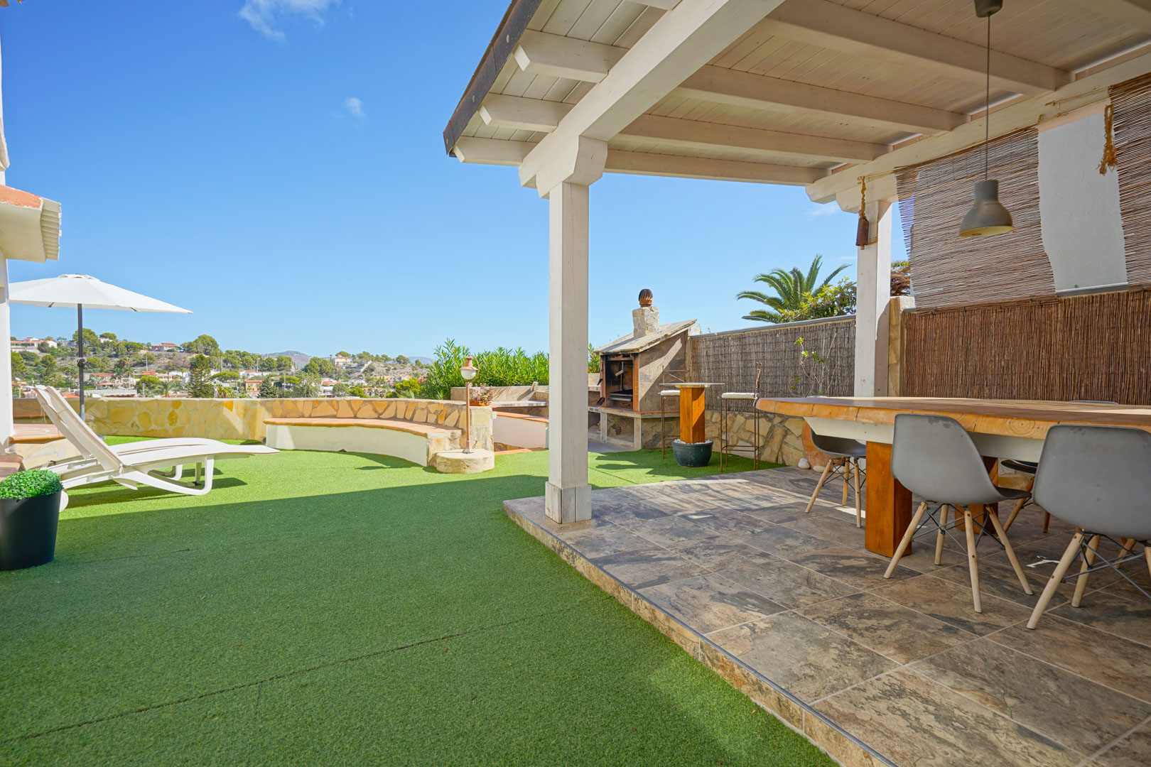 Terrasse extérieure avec pergola en bois blanc, coin repas, barbecue en pierre et espace en gazon synthétique avec chaises longues et vue dégagée.
