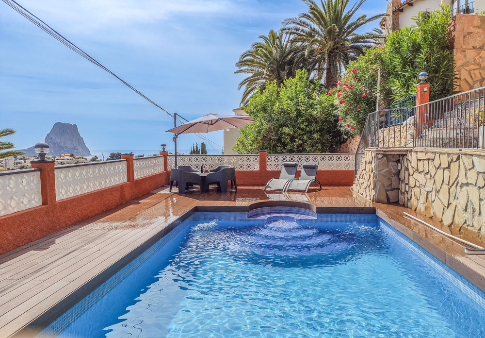 Terrasse extérieure avec piscine en mosaïque bleue, sol en bois et vue sur le Peñón de Ifach. Comprend un coin repas avec parasol et des chaises longues.