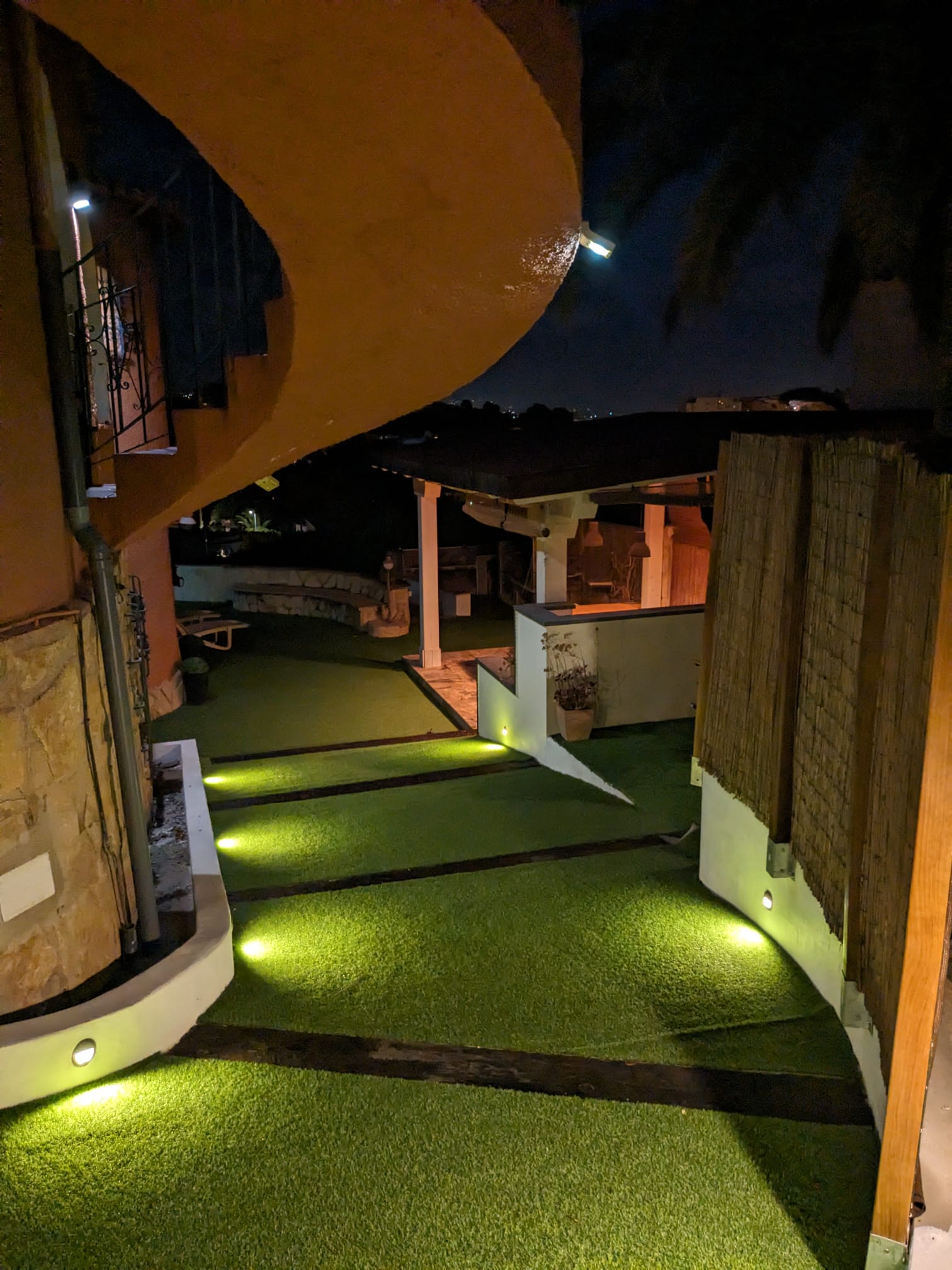 Vue nocturne des allées extérieures avec éclairage LED encastré, gazon synthétique et espace pergola couvert à l'arrière-plan.