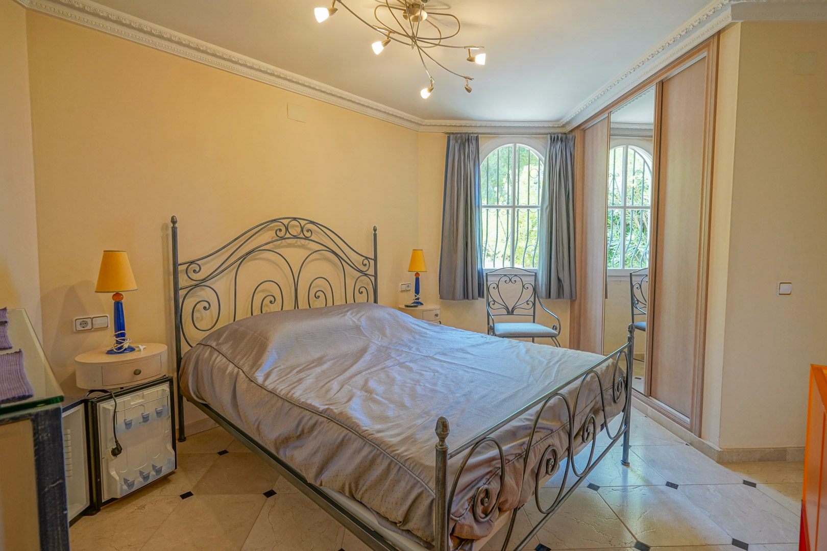 Chambre avec lit en fer forgé, sol en marbre à cabochons noirs, placards intégrés et fenêtre en arc offrant une vue sur la verdure environnante.