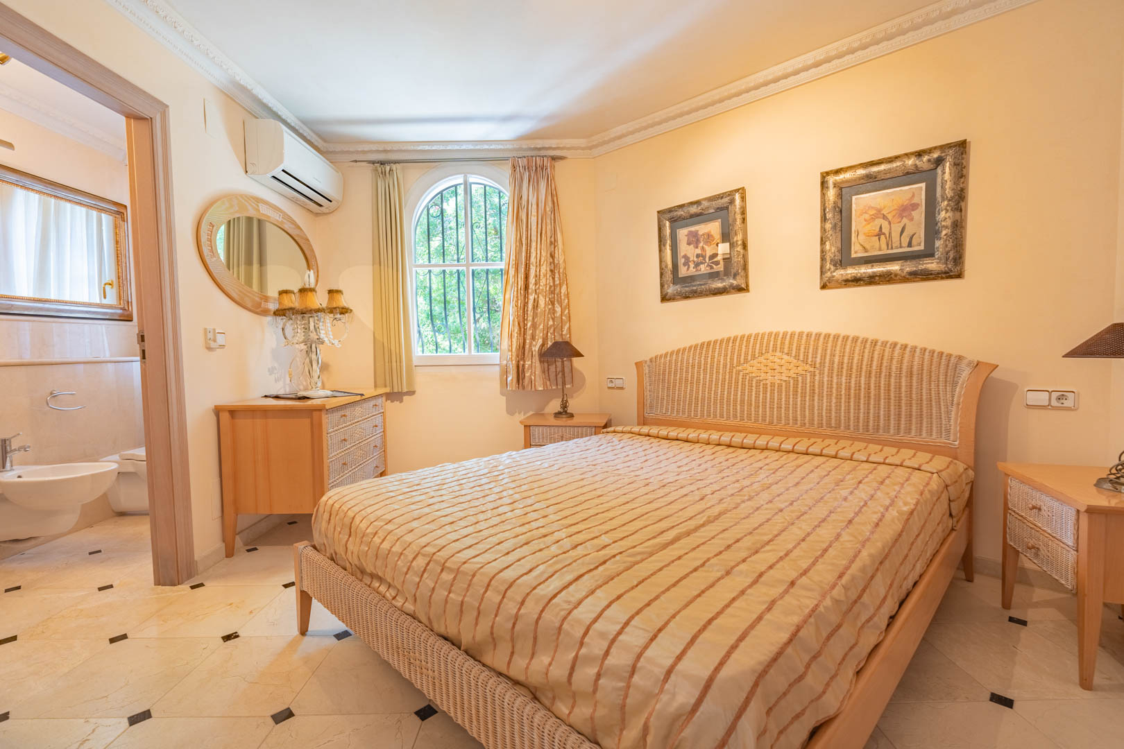 Chambre avec lit en rotin, sol en marbre à cabochons et salle de bain attenante. Elle dispose d'une fenêtre en arche, de moulures au plafond et de la climatisation.