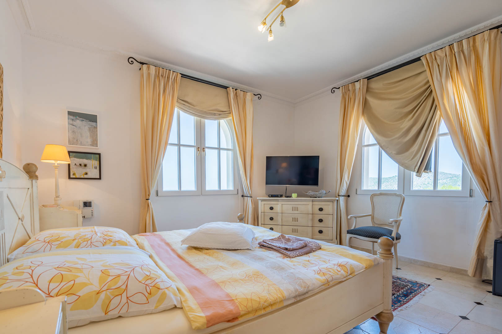 Chambre lumineuse avec mobilier en bois blanc, fenêtres cintrées aux rideaux crème et sol en carrelage avec cabochons. Comprend un coin télévision et un fauteuil classique.