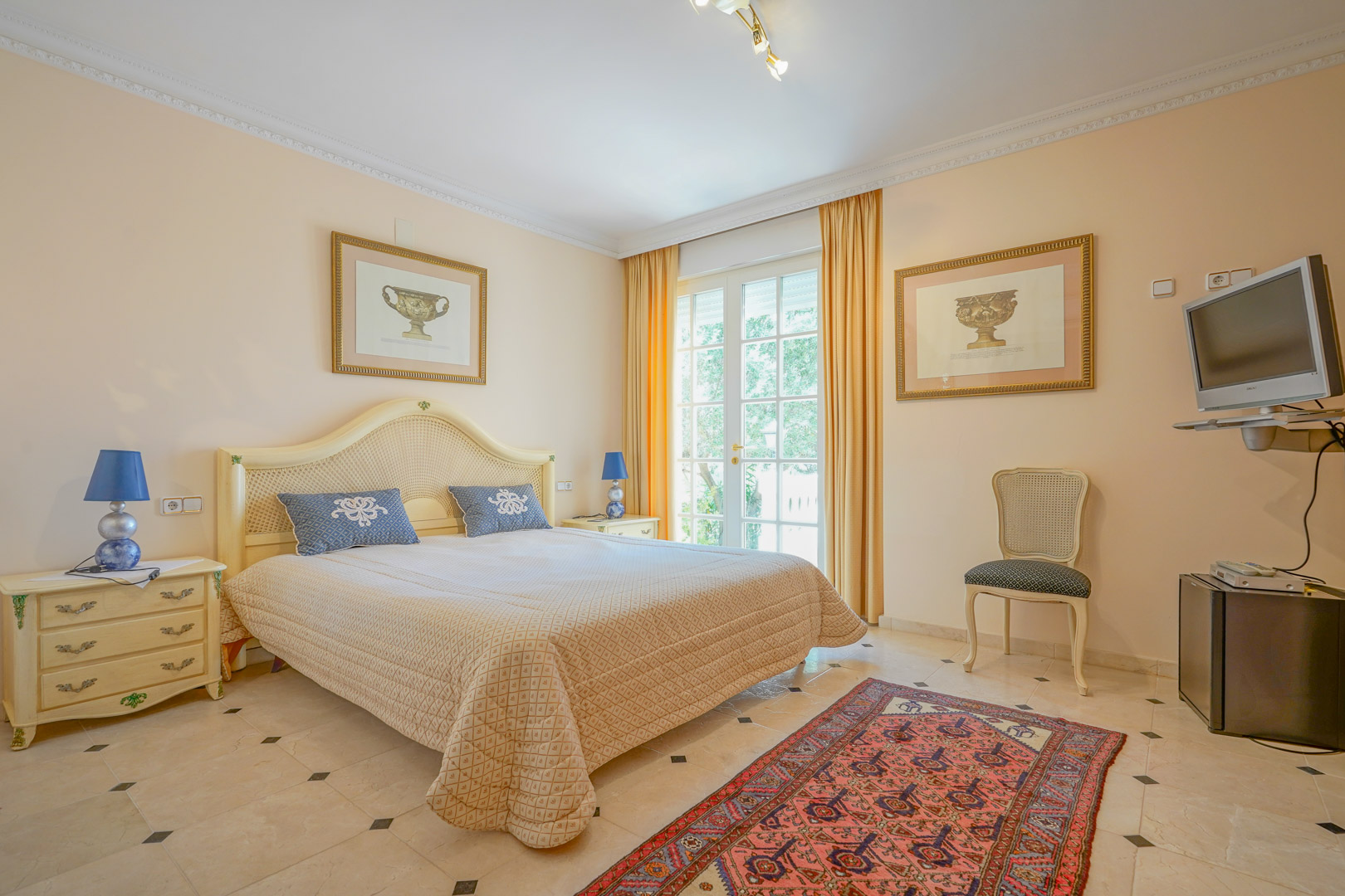 Chambre lumineuse avec sol en marbre et mobilier classique. Les portes-fenêtres offrent un accès direct à l'extérieur, complétées par une décoration aux tons crème et bleu.