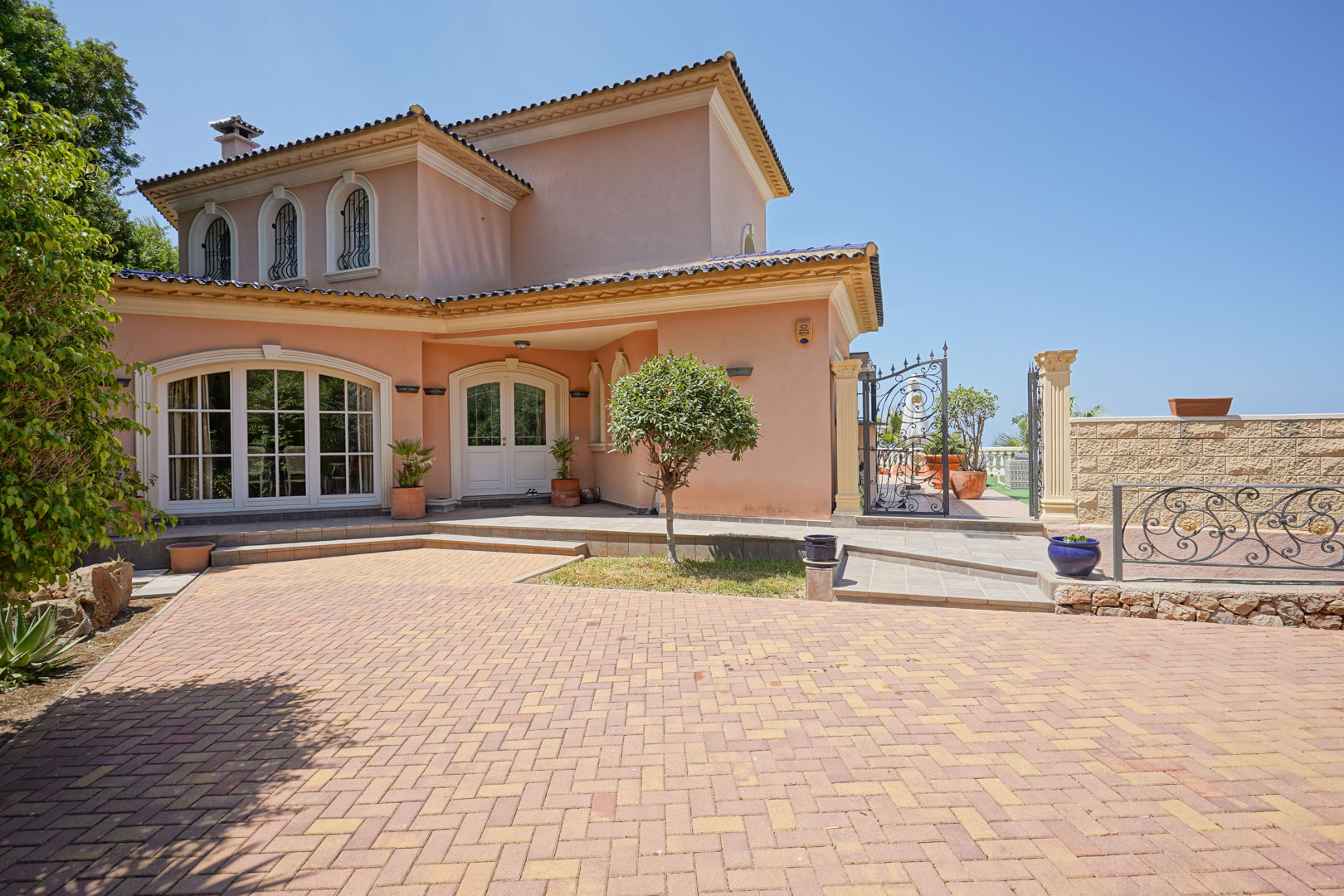 Façade de la villa avec entrée principale, fenêtres cintrées et large allée pavée. Détails en fer forgé et murs de couleur terre cuite sous un ciel azur.