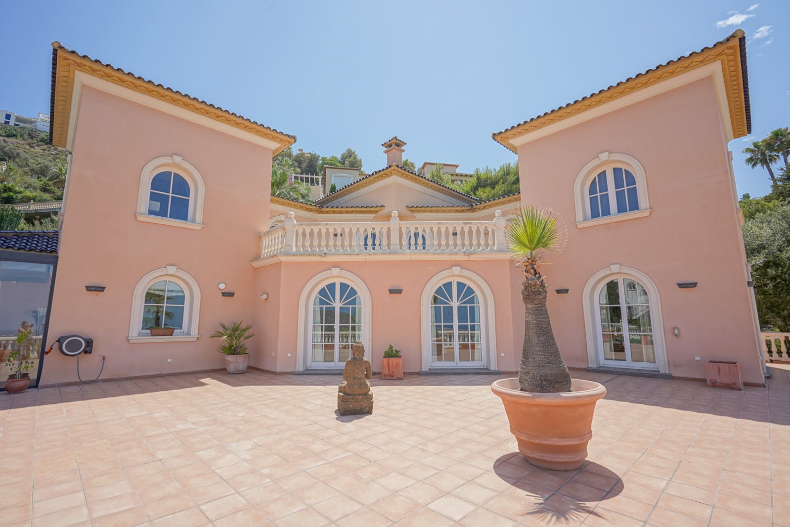 Façade symétrique aux tons saumon avec fenêtres cintrées, terrasse supérieure à balustrade blanche et vaste patio en terre cuite orné d'une statue en pierre.
