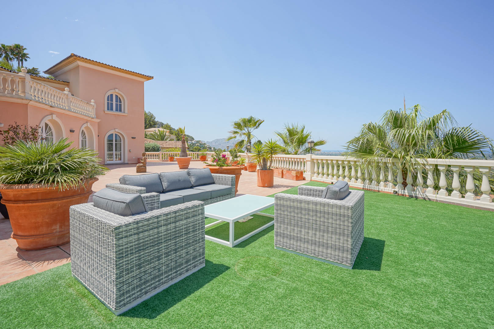 Grande terrasse avec salon de jardin en résine tressée grise sur gazon synthétique, offrant une vue panoramique sur la côte et les montagnes environnantes.