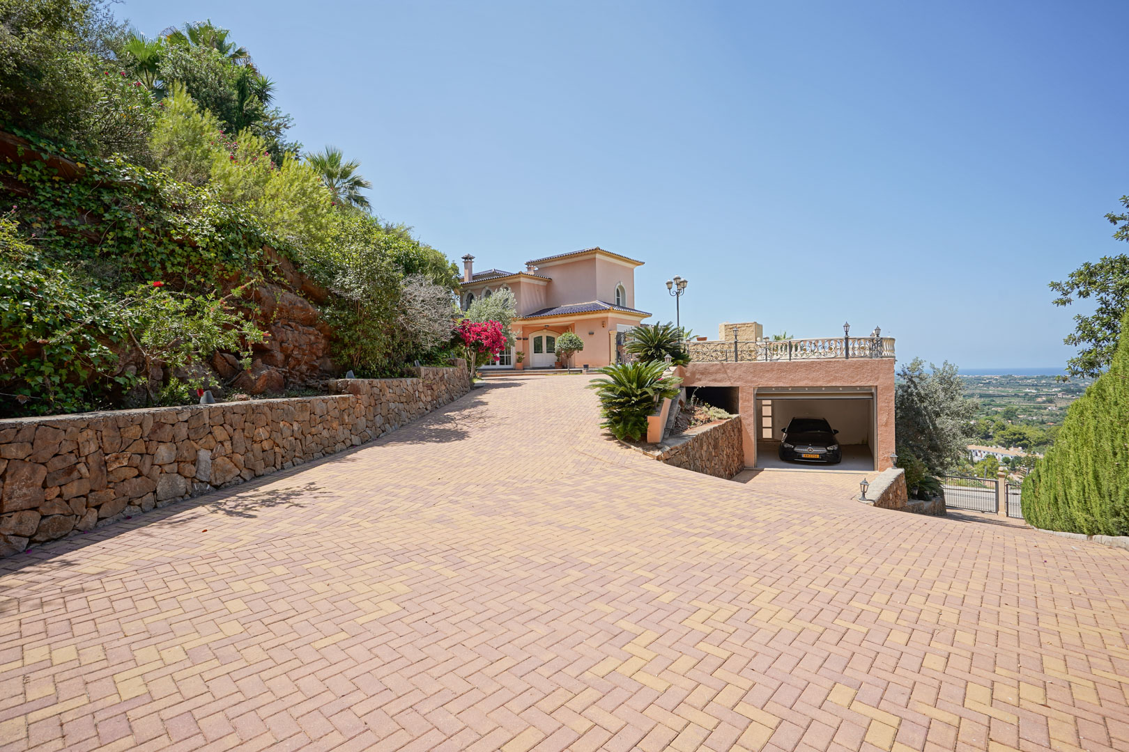 Large allée pavée menant à la villa et au garage intégré, bordée de murs en pierre naturelle et offrant une vue dégagée sur la vallée et la mer Méditerranée.