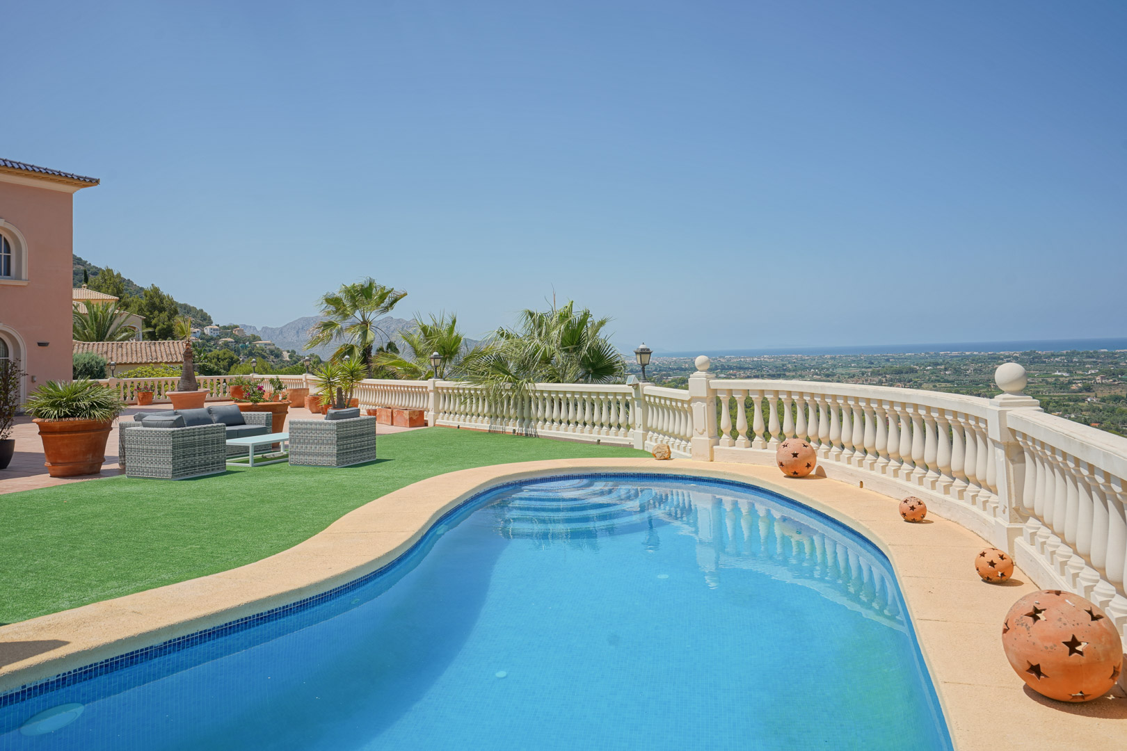 Piscine aux formes arrondies offrant une vue panoramique sur la mer, entourée d'une balustrade en pierre, de gazon synthétique et d'un salon de jardin élégant.
