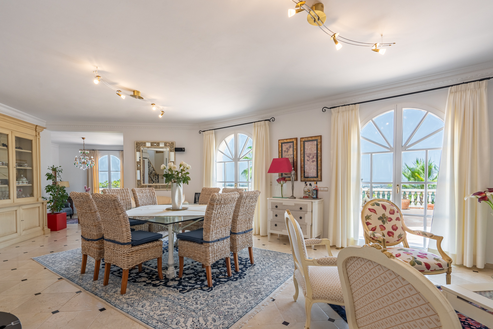 Salle à manger lumineuse avec chaises en osier et table en verre, s'ouvrant sur la terrasse par de grandes fenêtres en plein cintre.