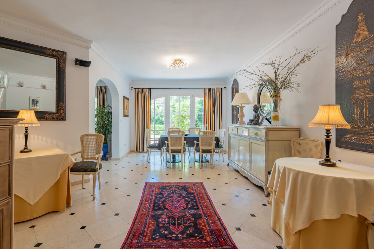 Salle à manger spacieuse avec sol en marbre et cabochons noirs, mobilier classique et grandes portes-fenêtres cintrées ouvrant sur le jardin verdoyant.