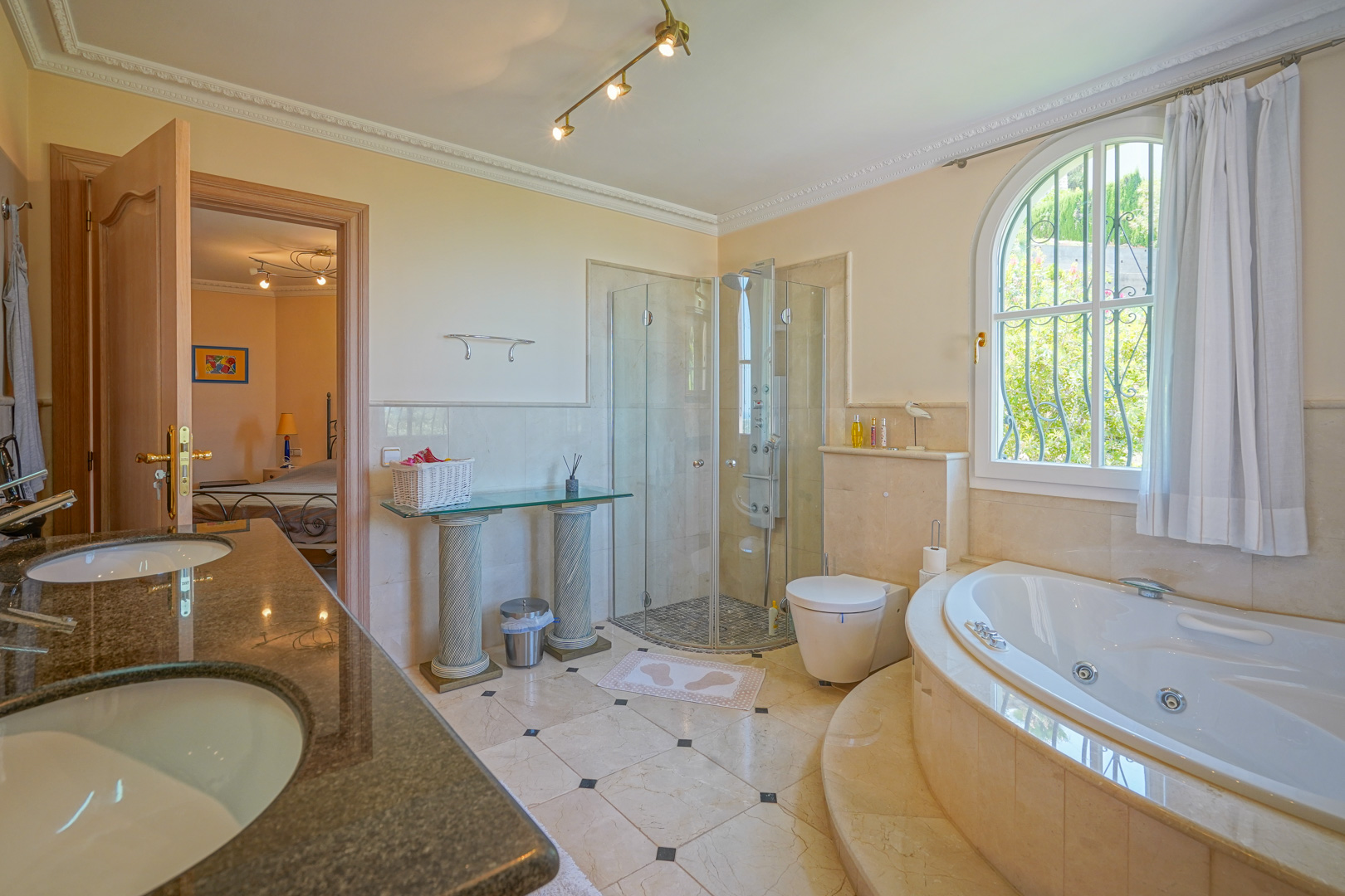 Salle de bain attenante avec baignoire d'hydromassage circulaire, douche vitrée et double vasque en granit. Sols en marbre et fenêtre cintrée offrant une vue sur la végétation.