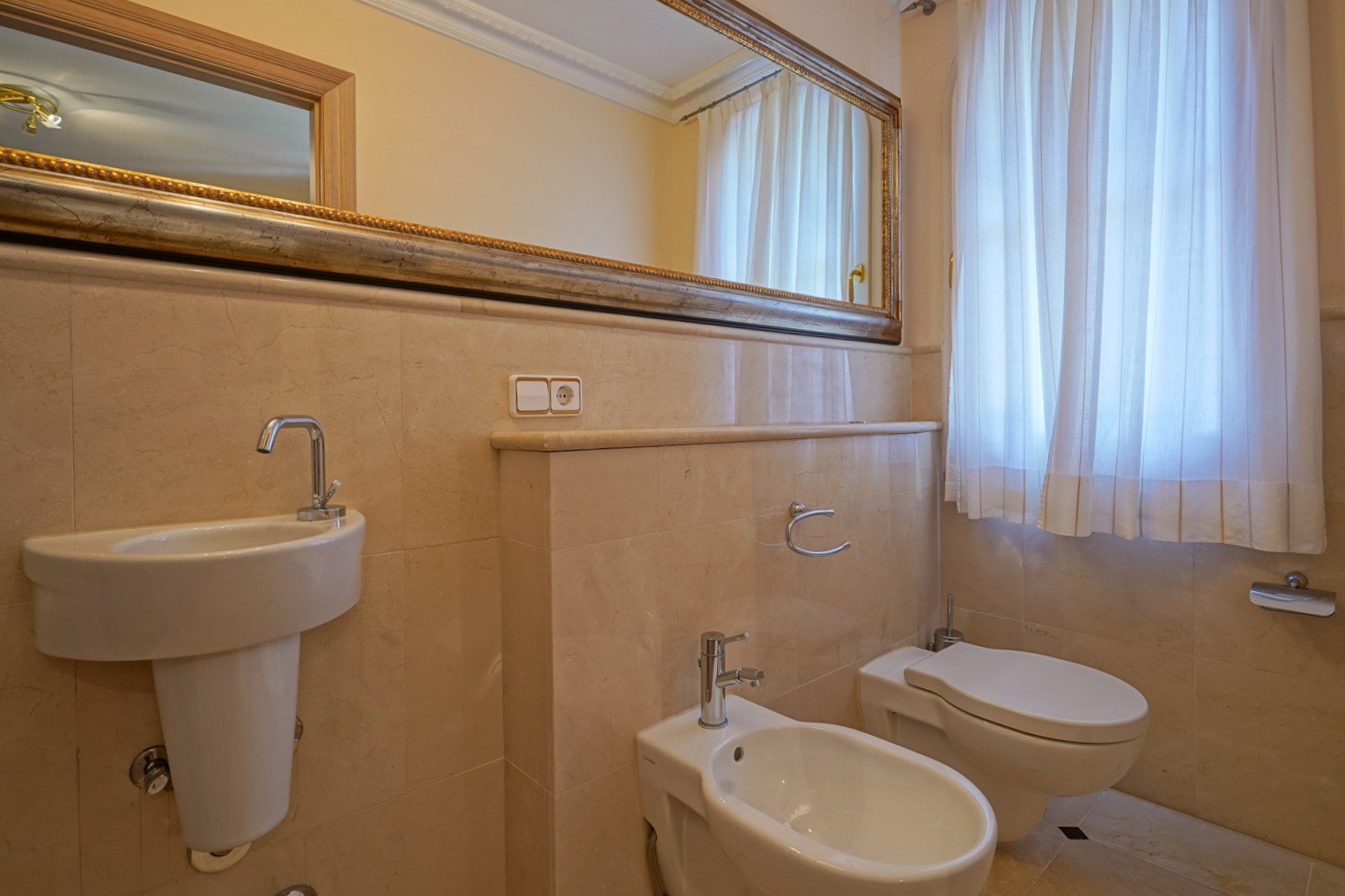 Salle d'eau avec revêtement en marbre crème, équipée d'un lavabo, d'un bidet et de toilettes. Un grand miroir à cadre orné et une fenêtre avec rideaux complètent l'espace.