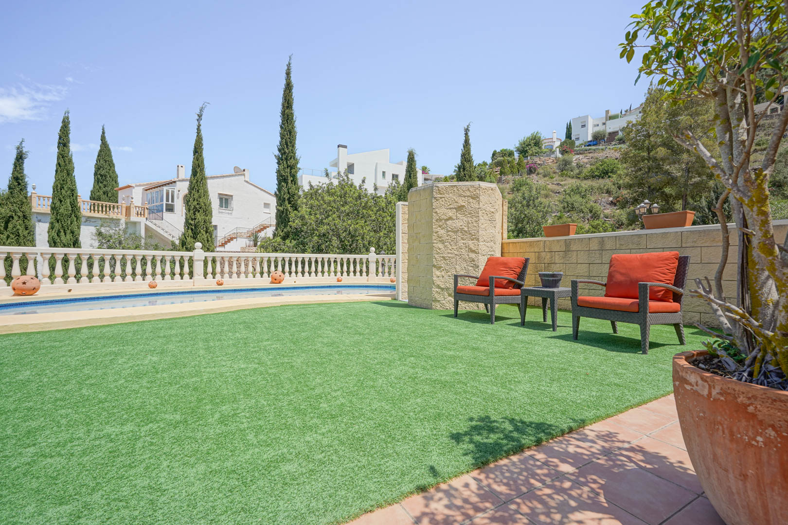 Terrasse extérieure avec gazon synthétique, coin salon aux fauteuils orange et piscine privée bordée d'une balustrade classique et de cyprès.