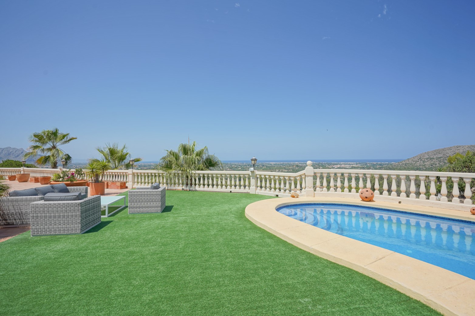 Terrasse extérieure avec piscine incurvée, gazon synthétique et salon de jardin en rotin offrant une vue panoramique sur la mer et la vallée de La Sella.