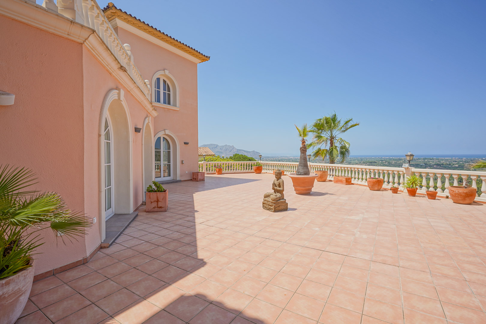 Vaste terrasse avec carrelage en terre cuite et balustrade offrant une vue panoramique sur la mer. La façade arbore des fenêtres cintrées et une décoration méditerranéenne.