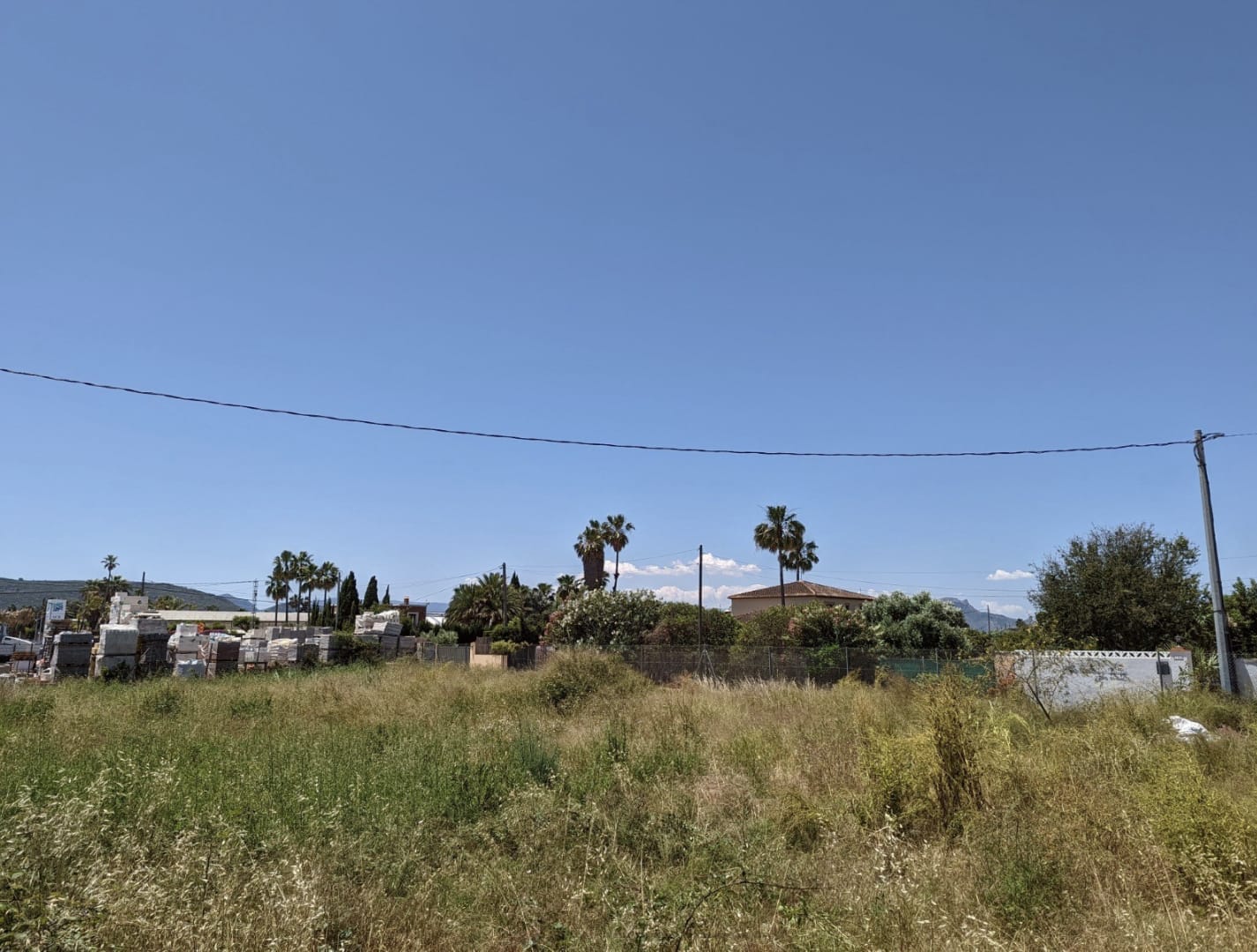 Terrain plat avec végétation basse et matériaux de construction entreposés. Vue dégagée sur des palmiers et des habitations environnantes sous un ciel bleu.