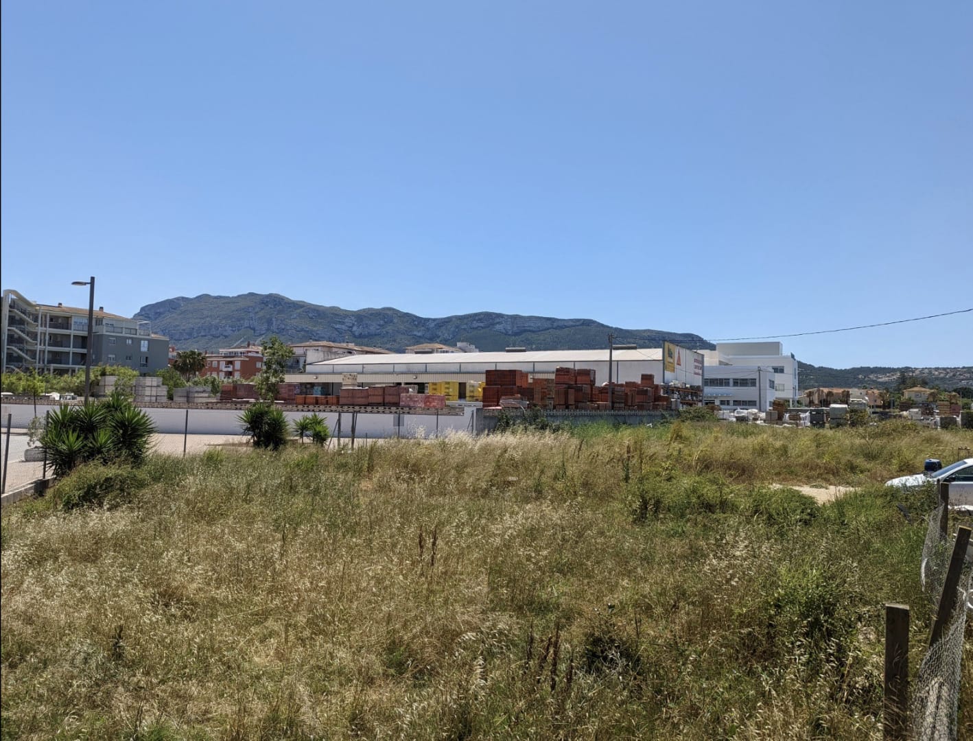Terrain plat avec végétation naturelle offrant une vue dégagée sur le Montgó, situé à proximité immédiate de zones commerciales et résidentielles.