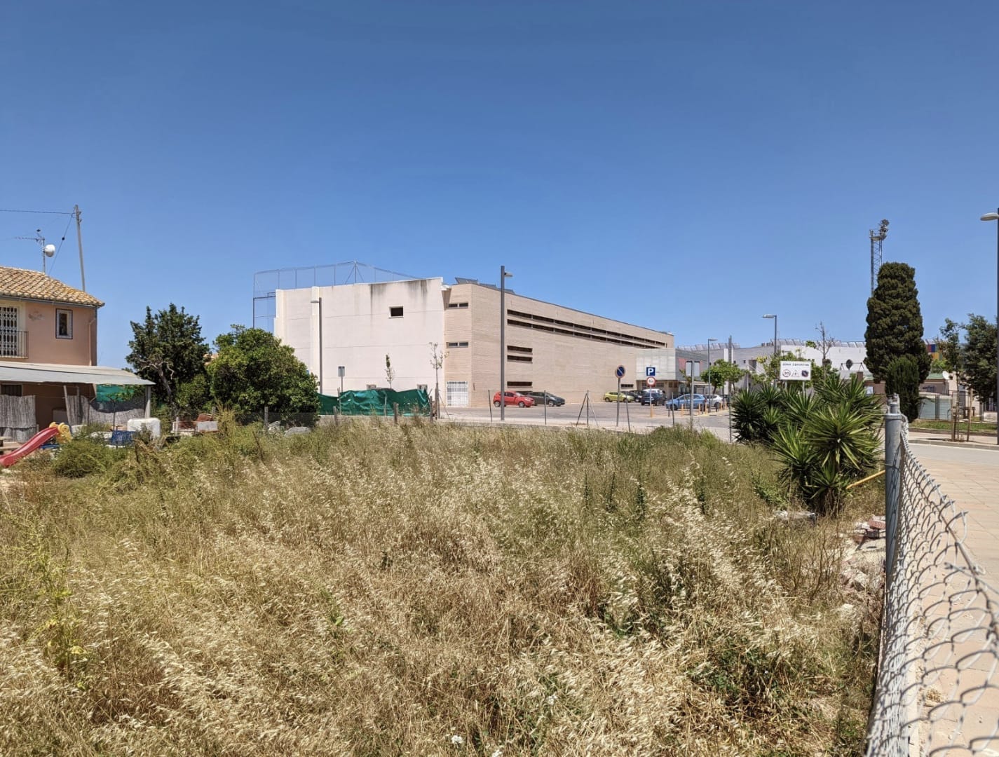 Terrain urbain plat avec végétation naturelle, situé face à un complexe sportif moderne et un parking public, sous un ciel dégagé.