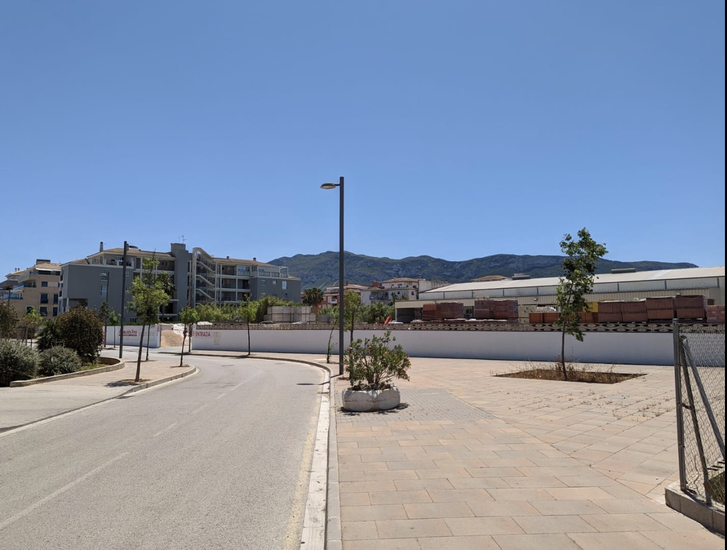 Vue du terrain urbain avec accès pavé et clôture périmétrale, offrant une vue sur le Montgó et la proximité des infrastructures résidentielles locales.