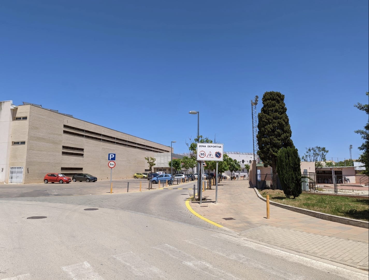 Vue extérieure de l'accès à la zone sportive de Denia, avec des trottoirs larges, une signalisation claire et des bâtiments institutionnels en briques.
