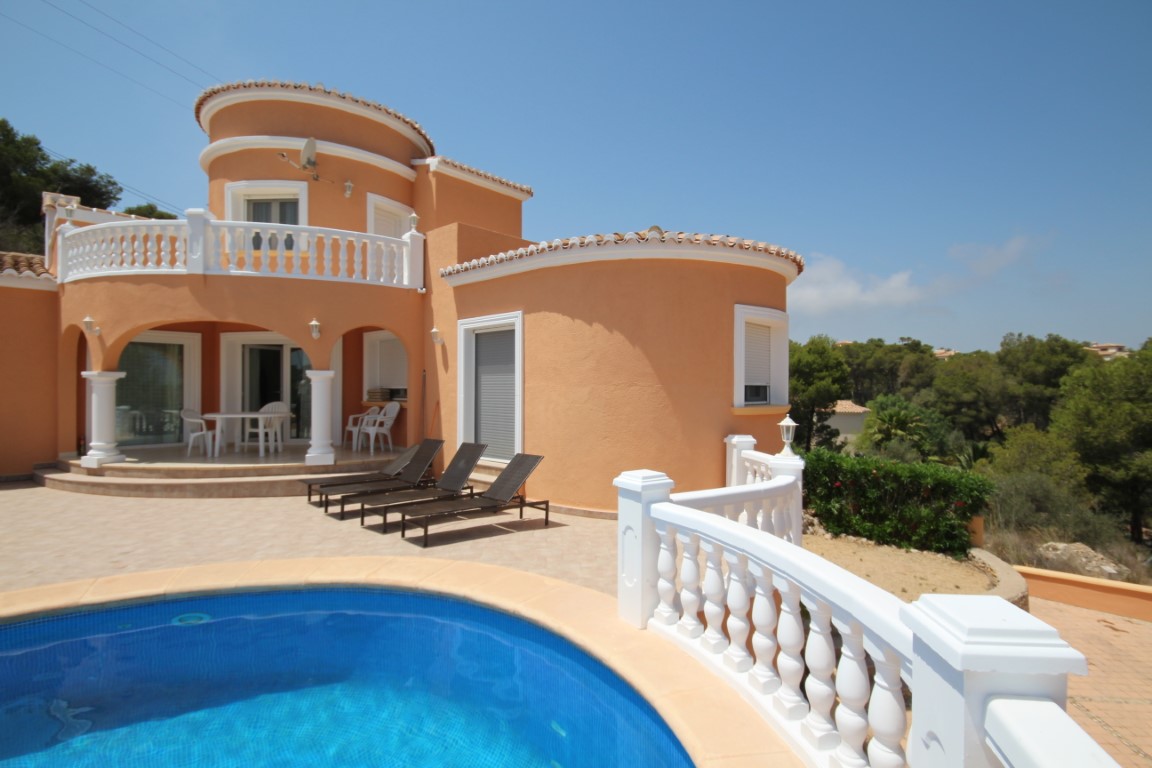 Extérieur de la villa avec piscine privée, terrasse ensoleillée et façade ocre. L'architecture se distingue par ses balustrades blanches et son porche couvert à arcades.