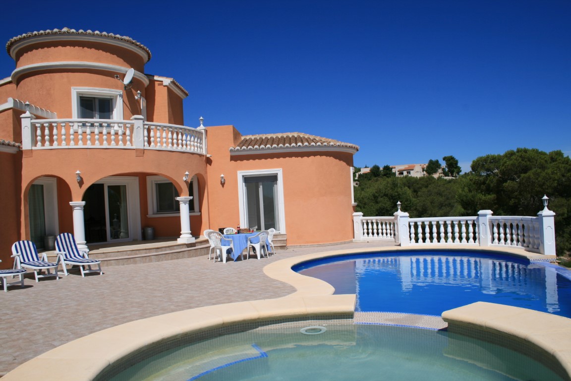 Extérieur d'une villa terre cuite avec piscine de forme libre et jacuzzi intégré, vaste terrasse carrelée bordée de balustrades blanches et arcades.